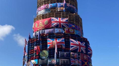 A large number of flags including union flags and a flag depicting an image of King Charles placed on a bonfire in Derry's Bogside.