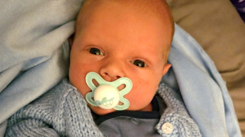 Baby Harley sucking a light blue dummy, wrapped in a blue fluffy blanket and baby grow.
