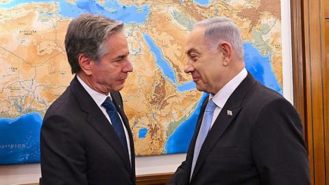 Blinken and Netanyahu shaking hands during a recent visit. The two men are standing front of a world map and look serious. 