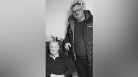 A black and white image of Ivor and Gregory McQuitty. Ivor is sitting in an armchair smiling as Gregory stands leaning onto the armchair also smiling. Ivor has short light hair and is wearing a jumper. Gregory has short light hair and a moustache, he is wearing glasses and a jumper with a coat over it. 