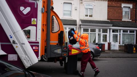 A photo of a person putting a black bin back into the back of a bin lorry. The person is wearing bright orange. 