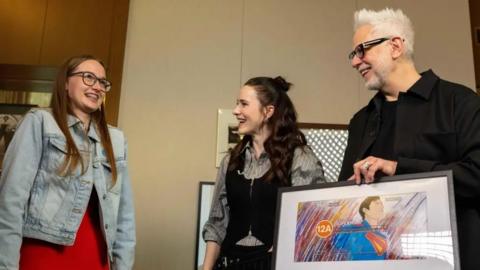 A girl in a denim jacket witha big smile as she talks to actor Rachel Brosnahan and director James Gunn in a room with soft, cream walls. James Gunn is holding a framed artwork depicting Superman in his blue superhero costume and an orange 12A age rating circle.