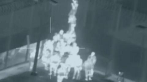 Heat-detecting footage from a helicopter shows a group of men scaling a fence.