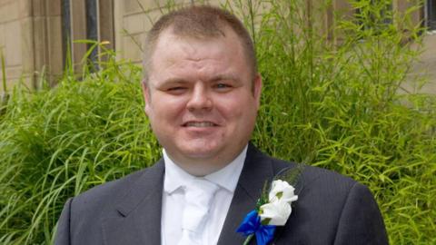 Neil Doyle standing in front of a green bush outside of a church, wearing a wedding suit with white flower in the buttonhole and smiling.