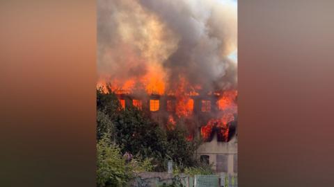 A burning building, with white and grey smoke in the air. Large flames can be seen coming from the building.