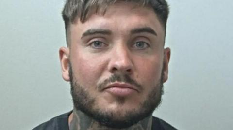 A police mugshot of Ryan Wellings who has short brown hair and a short brown beard, and a tattoo covering most of the front of his neck.