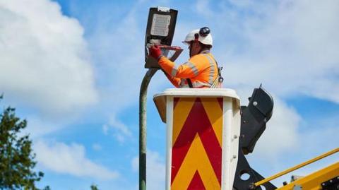 A contractor in a crane's cherry-picker fits an LED streetlight. He is wearing a white helmet and orange hi-viz jacket.