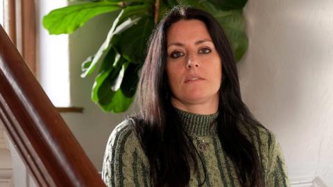 Rebecca has long dark hair and is wearing a green and cream polo neck sweater. She is sitting on the stairs with her hands on her lap and looking over the bannister into the camera. There is a plant with big green leaves behind her. She is not smiling.