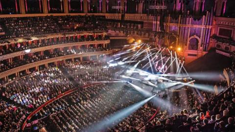 A full Royal Albert Hall from the top tier and looking down at a stage with lots of lights.