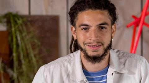 Tayshan Hayden-Smith is seen beard wearing a white jacket, smiling in front of a wooden wall and red geometric sculpture.