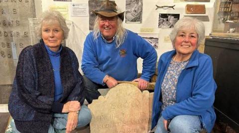 An elderly man in the middle and two elderly women either side with a gravestone in between them.