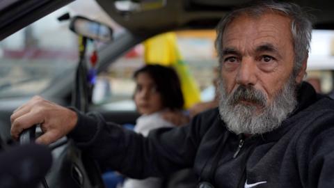 Abu Ali sits in the front seat of his car, hand on the steering wheel. A child and presumed relative sits in the passenger seat next to him.