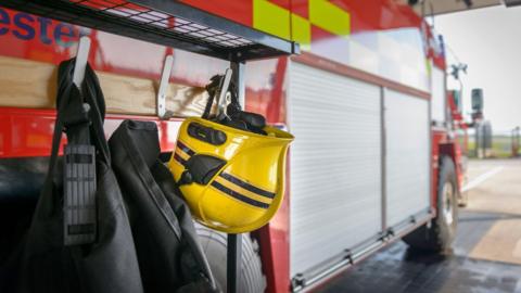 A photo of firefighting gear - a yellow firefighter helmet with black and reflective stripes - hanging on a hook on a fire engine. The fire truck is parked inside.