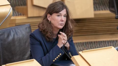 Dorothy Bain, who has brown curly hair, sits with her hands clasped in front of her at a desk in the Scottish Parliament. She is wearing a navy blue jacket.