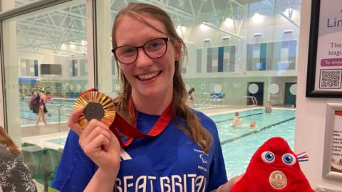 Becky Redfern holds up her gold medal in one hand and a Paris 2024 mascot in the other. She is standing in front of a window overlooking the swimming pool.