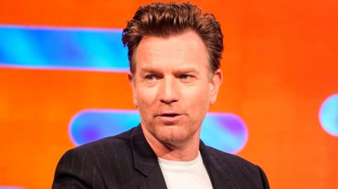 A man, Ewan McGregor, speaks in front of a bright orange background - he has short brown hair and wears a black jacket