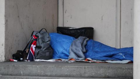A homeless person sleeping rough in a doorway. Their face and body is covered by a light blue sleeping bag and a grey rucksack blocks view of the person's head.