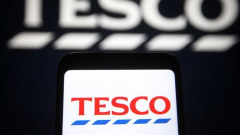 The Tesco logo is displayed on a smartphone, against a black background displaying a larger, blurred version of the supermarket's logo.