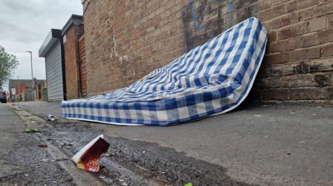 Blue and white mattress and some litter in an alleyway in front of a brick building.