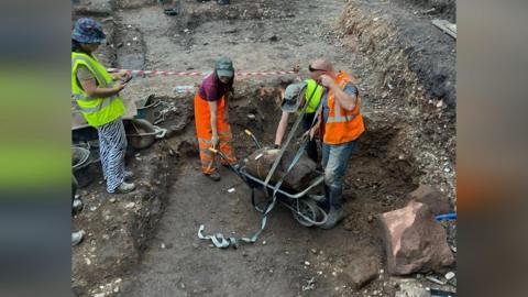 Three volunteers wearing high viz clothing secure the section of column to a wheelbarrow while another is stood next to them taking a picture on her phone. They are in the middle of a dug-up area.