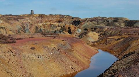 A rusty looking mountain side bearing the scars of centuries of mining. Its lunar landscape is bereft of vegetation. There is a large puddle of water in the foreground of the shot. In the background you can see a pumphouse which removed water from the underground tunnels