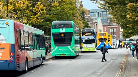 A row of buses are parked at a variety of bus stops on a street. To the right a number of people are walking on a pavement. There are buildings in the distance and trees on the left and right hand side.