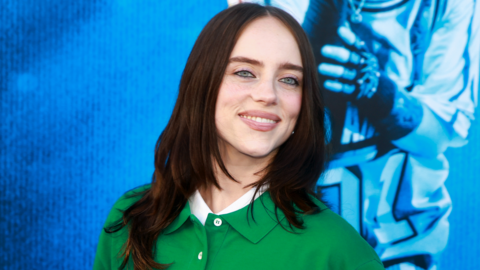 Billie Eilish smiling at the cameria wearing a green cardigan against a blue background