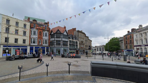 A wide pedestrian square in Wolverhampton city centre, with historic buildings lining the edges, bunting strung overhead and people walking across open paving. Shops and seating areas surround the square under a cloudy sky.