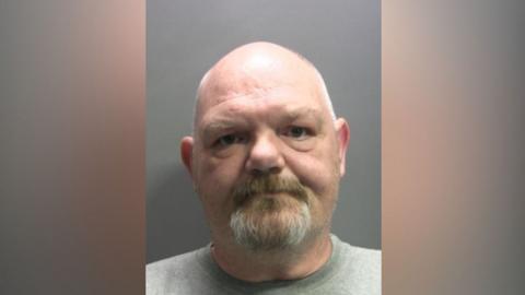 A mug shot of Stuart Sanderson. He has a grey and brown goatee beard and a bald head.