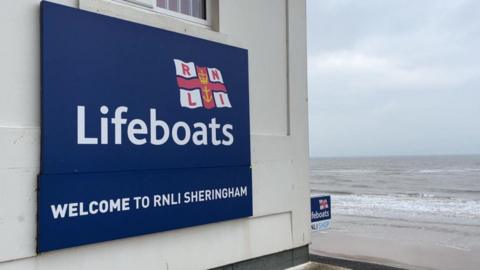 A white lifeboat station building. There is a blue sign on the side of the building with white writing which reads, "Lifeboats WELCOME TO RNLI SHERINGHAM". The sea is in the distance, with the lifeboat station next to the beach.