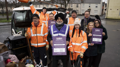 Street cleaners and neighbourhood co-ordinators stand with councillor Bhupinder Gakhal (centre) next to a pile of rubbish by a street bin in the city. They have florescent jackets on and some are holding poster about the scehem.