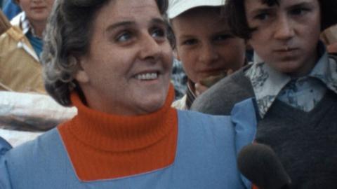 Eleanor, wearing an orange poloneck and blue apron, speaks to the reporter