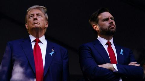 Donald Trump and JD Vance stand side-by-side, wearing navy suits and red ties. Trump has fair hair and has a serious expression on his face. Vance has brown hair and a beard, and looks quizzical. 