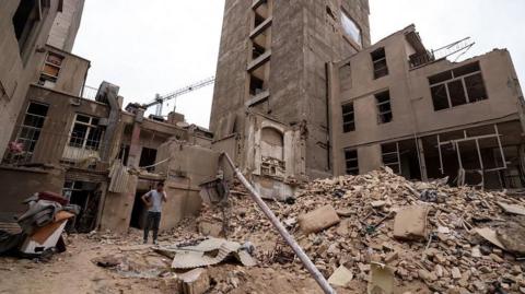 An Iranian man speaks on his mobile phone as he stands on the debris in front of destroyed buildings following a military strike on the Iranian capital Tehran
