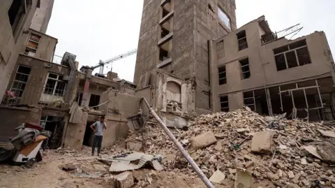 An Iranian man speaks on his mobile phone as he stands on the debris in front of destroyed buildings following a military strike on the Iranian capital Tehran