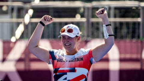 Phoebe Paterson Pine is wearing a red, white and blue t-shirt that says "Great Britain". She has her arms in the air, celebrating. She is smiling and wearing a white cap, with white sunglasses resting on it