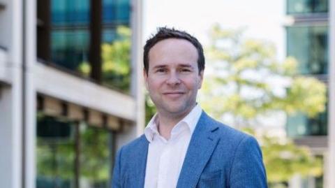 Paul Wylie smiling at the camera and stood in front of buildings. He is wearing a white shirt and a blue blazer and has his hands in his pockets.