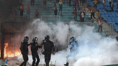 Riot police, tear gas and a fire all in front of a stand with spectators