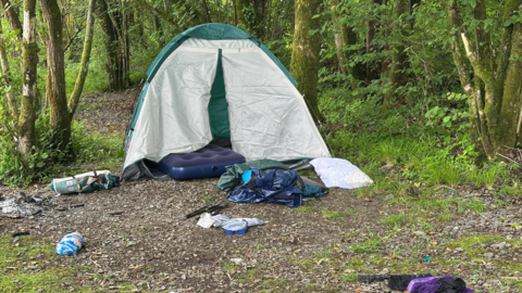 A tent surrounded by litter in picturesque woodland in the Lake District. There is a lot of rubbish and discarded camping equipment. 
