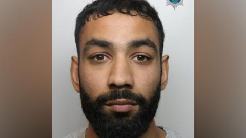 A custody image of Zohaib Mehmood. He has black hair and a black beard and is looking at the camera. 