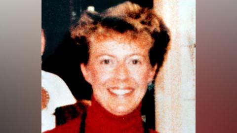 An old photo of Janice Cartwright-Gilbert is wearing a red polo-neck jumper. She has short blonde hair and is smiling broadly at the camera. 
