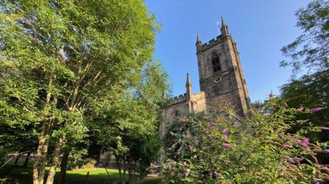 The tower of a church building is seen, with lower parts of the structure obscured by bushes in the foreground.