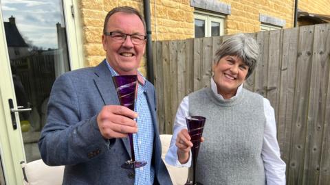 Colin Durber is shown on the left wearing a blue suit jacket and black rimmed glasses. Corinne Durber is on the right and wears a white long-sleeved shirt and a grey sweater vest. They are both raising purple champagne flutes in their hands.