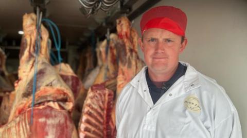 A man in a large white coat and a red hat stood in front of butchered meat hanging from a ceiling