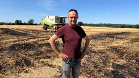 A man standing in a field where the harvest has been destroyed by a fire. Parts of the ground are black. The man is wearing light blue jeans and a burgundy top. He has his hands on his hips. The sky is blue in the background.