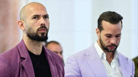 Andrew Tate, wearing a black top and purple blazer, stands next to his brother Tristan Tate, wearing a white shirt and light purple blazer