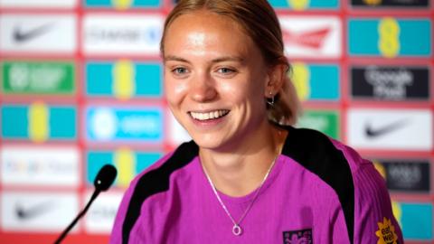 Esme Morgan smiles as she sits in front of a microphone wearing a pink England training top. She has blonde hair tied back in a pony tail, a silver hoop earing in her left ear and a silver necklace. She is sat in front of a blurry background featuring various sponsorship logos.
