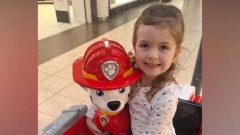 A young girl with brown hair and a white top. She is in a toy car featuring a character from Puppy Patrol.