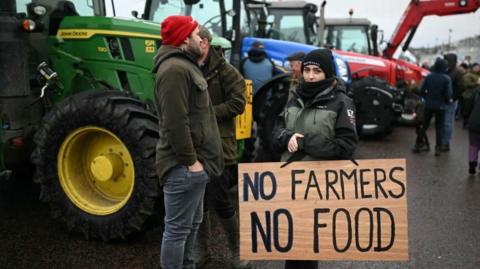 A woman wearing a green and black jacket and a black wooly hat holds a sign that reads "No Farmers, No Food". Behind her are stood a number of other farmers, as well as tractors lined up. 