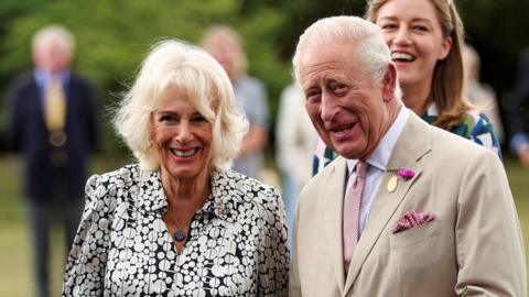 King Charles and Queen Camilla smiles while looking away from the camera.
